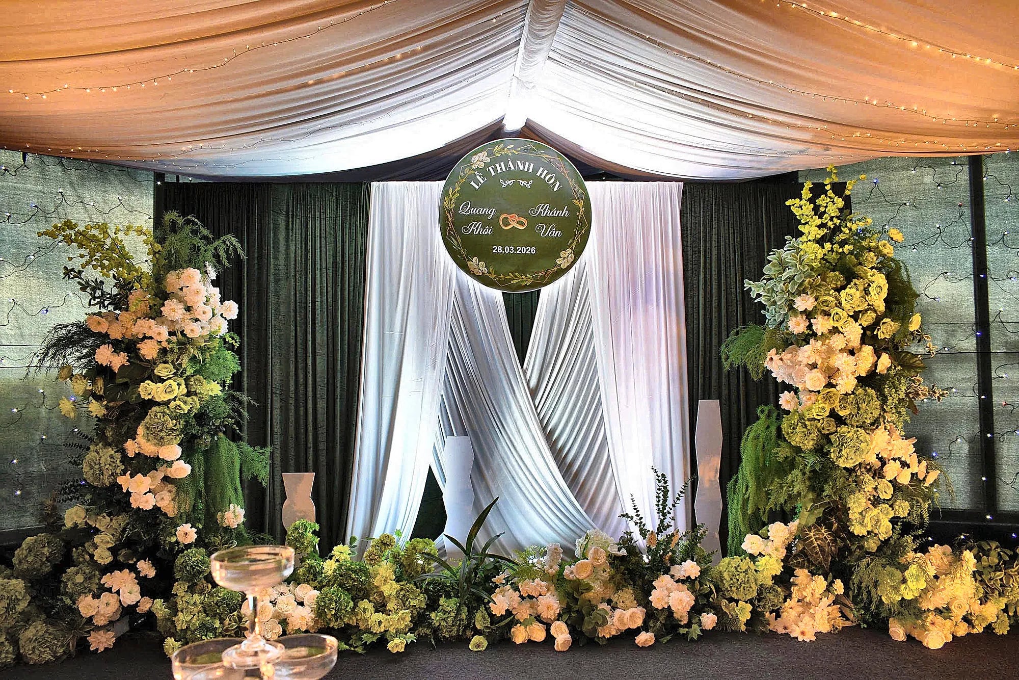 Backdrop tiệc cưới với rèm trắng, hoa xanh trắng và bảng tên lễ thành hôn